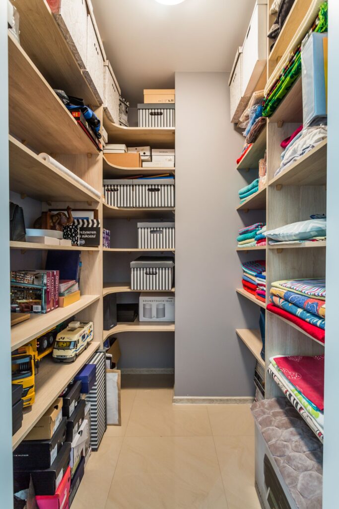 Pomieszczenie z drewnianymi półkami, pudłami, książkami, zabawkami i kolorowymi tekstyliami. Wnętrze jest posprzątane i uporządkowane.