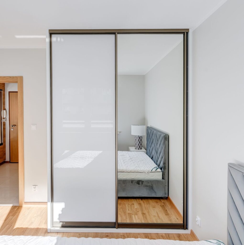 Lustro na drzwiach szafy odbija fragment sypialni z łóżkiem i lampką na stoliku nocnym, drewniana podłoga.