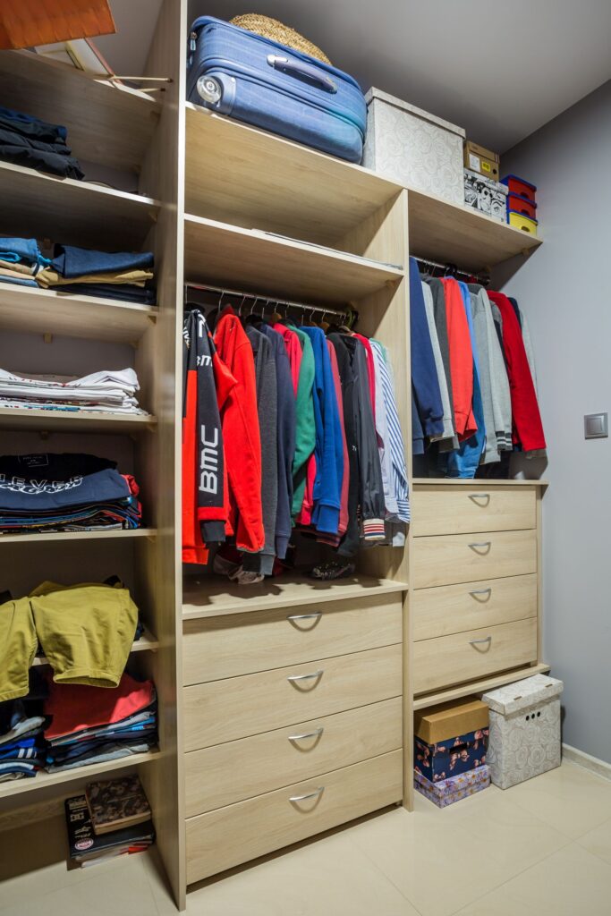Szafa z drewnianymi półkami, kolorowymi ubraniami, walizką i pudełkami na górze, uporządkowana przestrzeń.