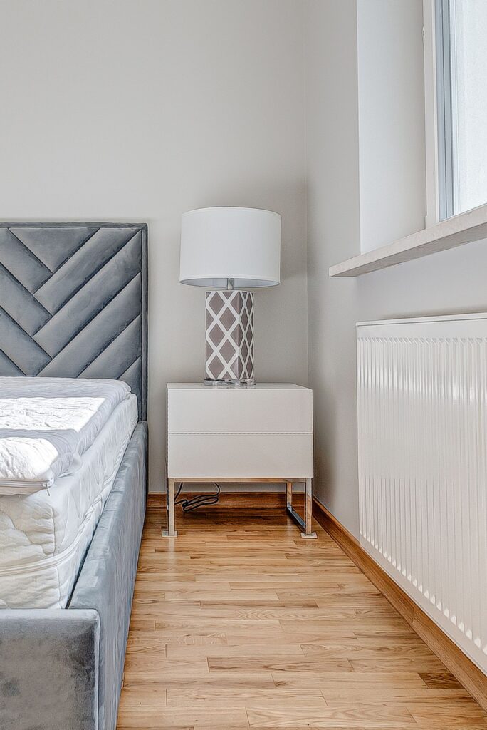 Nowoczesna sypialnia z szarym łóżkiem, białą szafką nocną i lampą z geometrycznym wzorem. Jasne, minimalistyczne wnętrze.