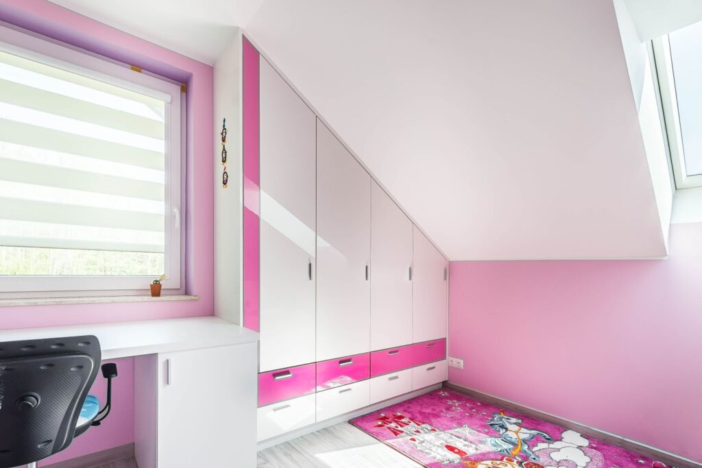 Pokój dziecięcy w różowych odcieniach z szafami, biurkiem i kolorowym dywanem. Światło wpada przez okno dachowe.