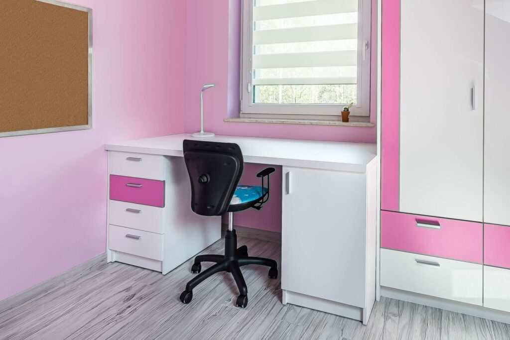 Biurko w różowym pokoju z krzesłem, białą lampką, tablicą korkową i szafką przy oknie z roletą.