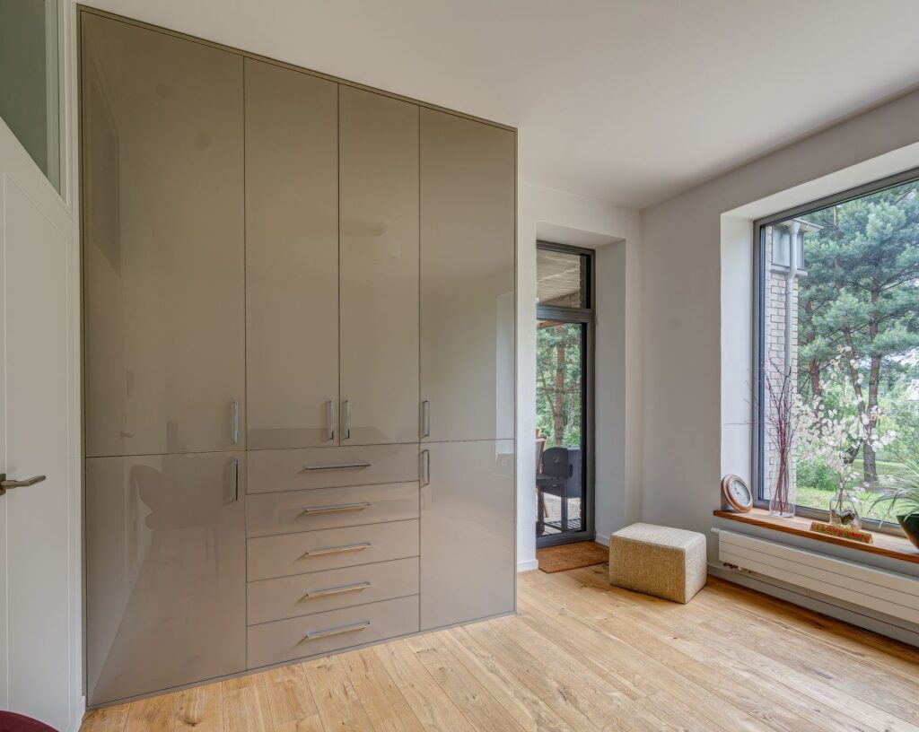Pokój z beżową szafą, dużym oknem z widokiem na drzewa, jasną podłogą i pufą przy ścianie.
