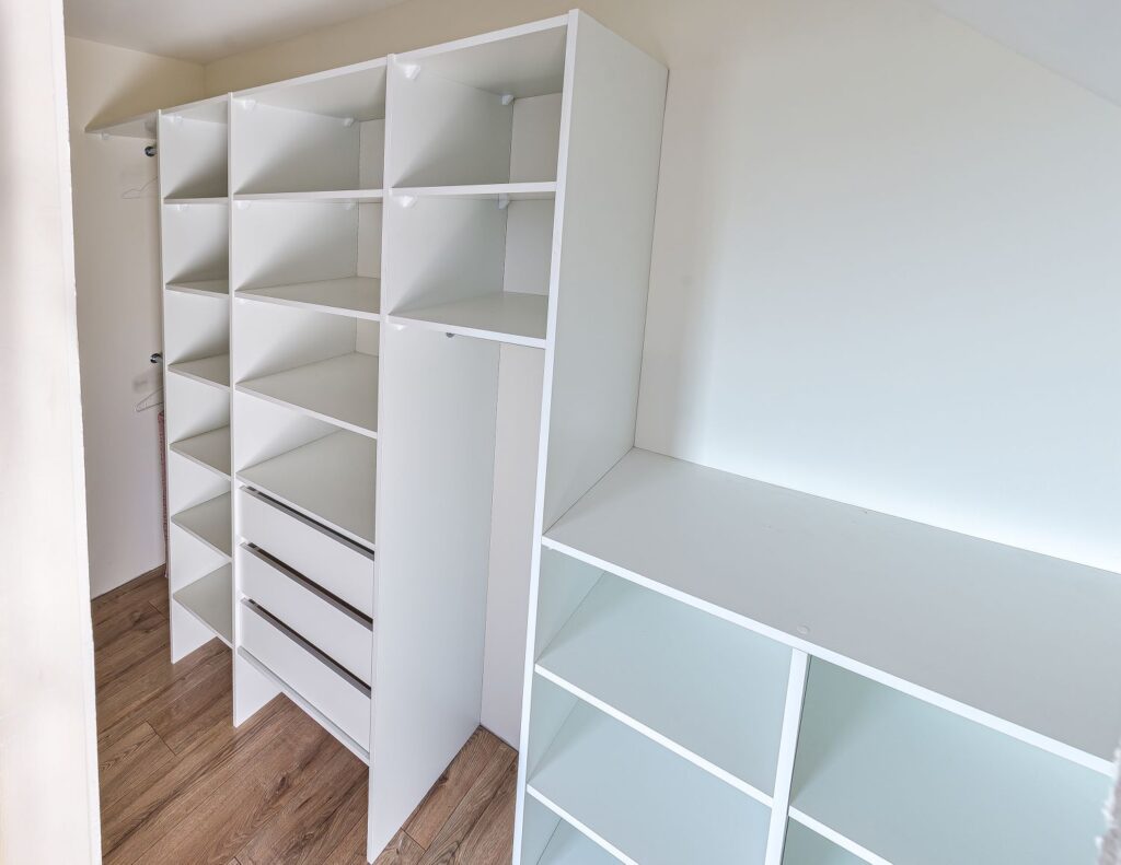 Biała szafa wnękowa z szufladami i półkami, na tle drewnianej podłogi.