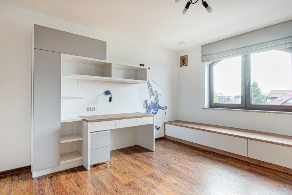 Biurko z półkami w jasnym pokoju, na ścianie grafika piłkarza, obok duże okno z widokiem na dachy i drzewa.