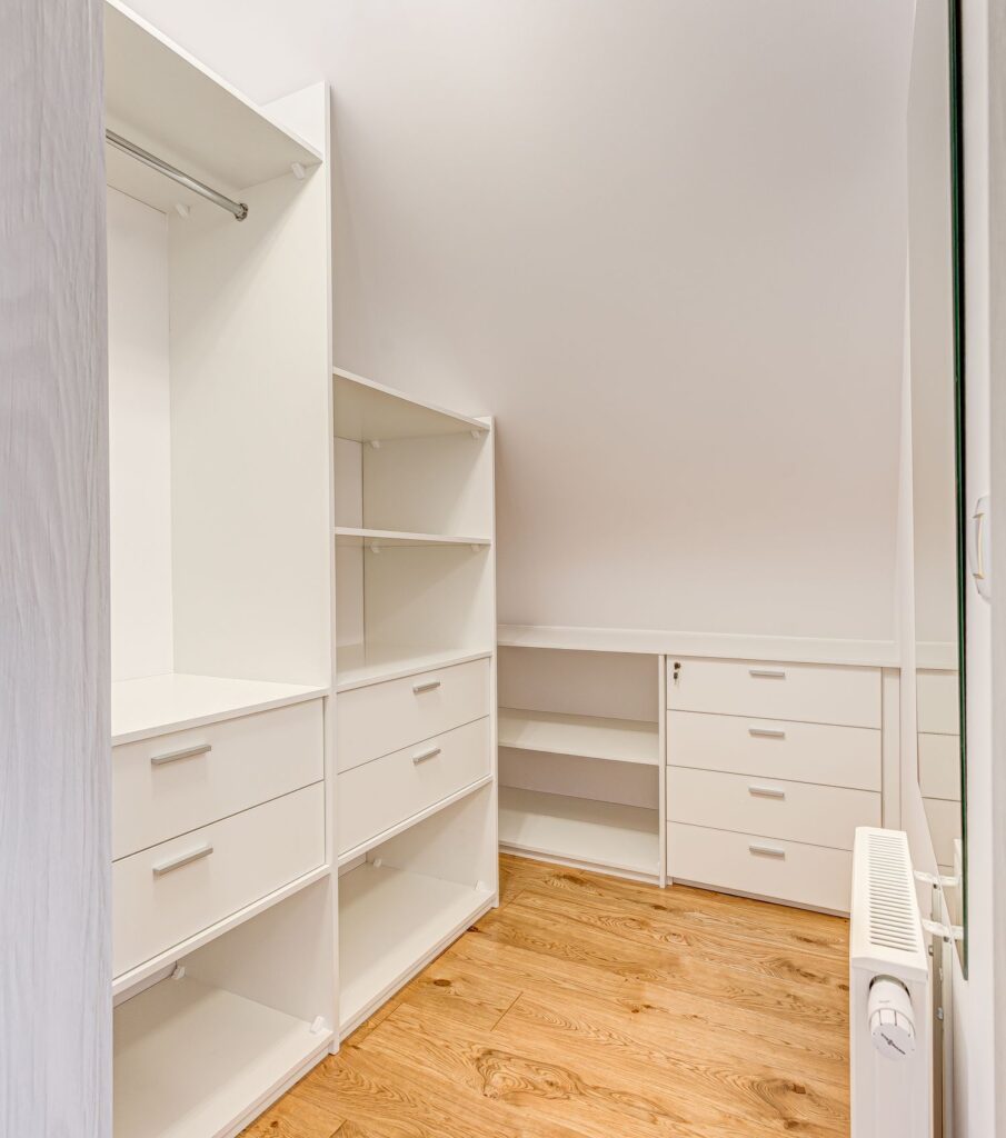 Biała, przestronna garderoba z otwartymi półkami i szufladami, drewniana podłoga, skośny sufit, kaloryfer po prawej stronie.