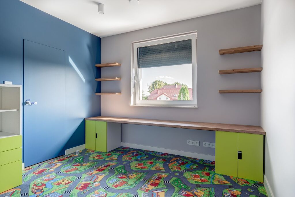 Pokój z niebieską ścianą, kolorowym dywanem z motywem drogi, półkami i dużym biurkiem. Widok na domy przez okno.