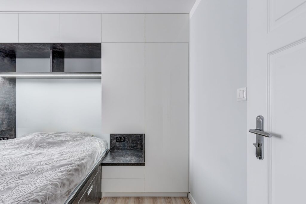 Nowoczesna sypialnia z białymi meblami, szarymi detalami i dużym łóżkiem, widok na drzwi z uchwytem.