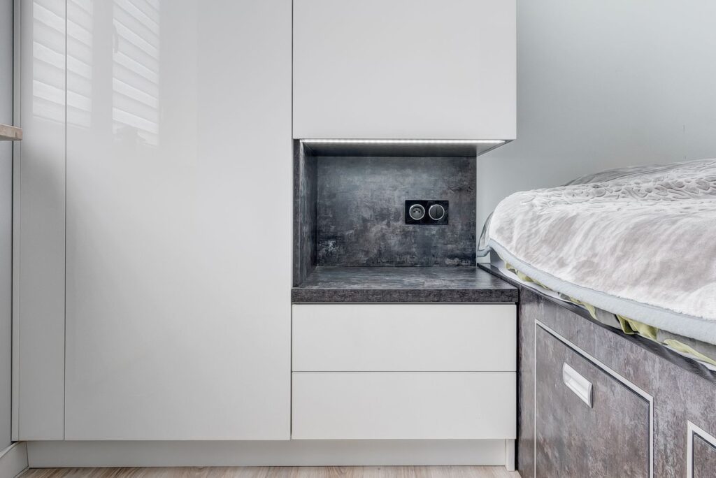 Biała szafka z gniazdkiem elektrycznym obok łóżka w nowoczesnym wnętrzu.
