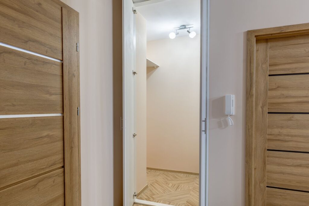 Otwarta szafa z białymi drzwiami, oświetlona lampka. Obok drewniane drzwi i domofon na ścianie.