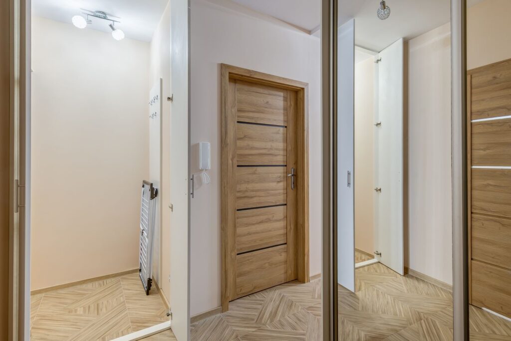 Wnętrze korytarza z drewnianymi drzwiami i lustrzaną szafą, jasne ściany i nowoczesne oświetlenie.