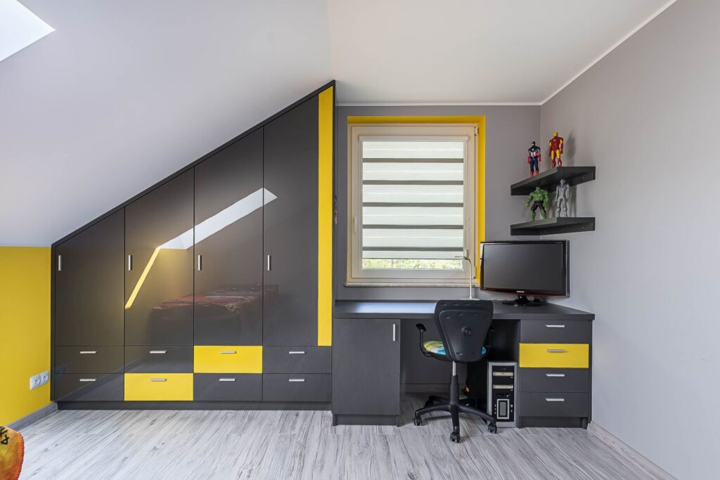 Pokój na poddaszu z biurkiem, komputerem i figurkami. Szafa czarno-żółta, okno z roletą, jasna podłoga.