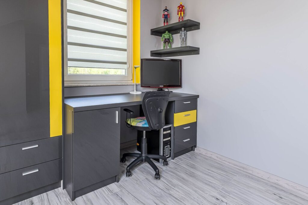 Nowoczesne biurko w pokoju z szarymi i żółtymi akcentami, figurki superbohaterów na półkach, komputer i krzesło obrotowe.