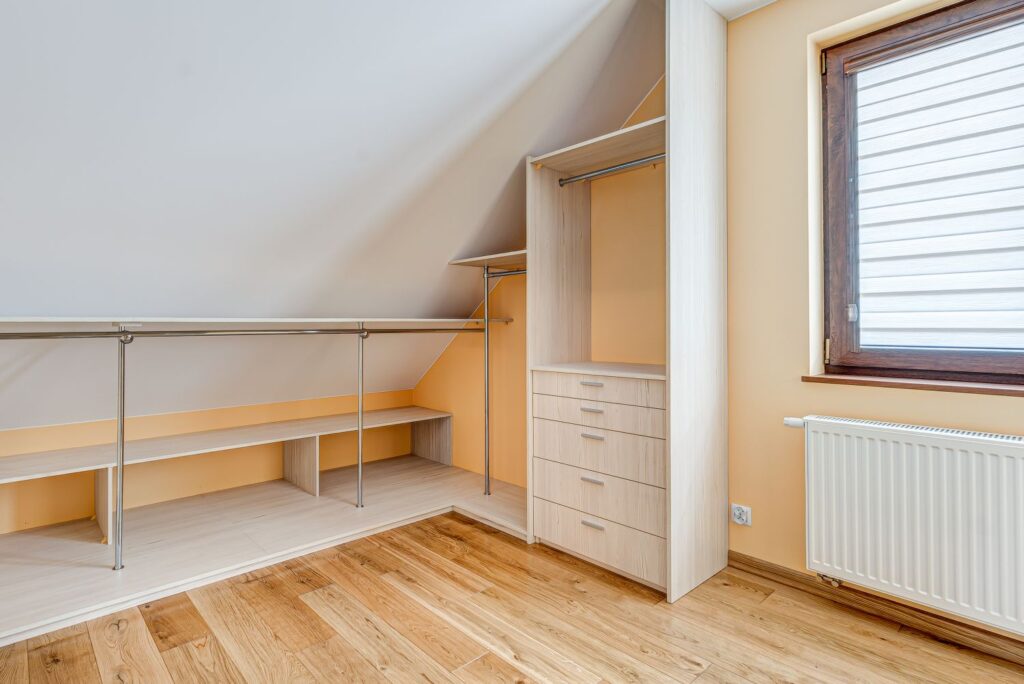 Pusty pokój z szafą wnękową, wieszakami, półkami i dużym oknem; jasnobrązowe ściany i podłoga z drewna.