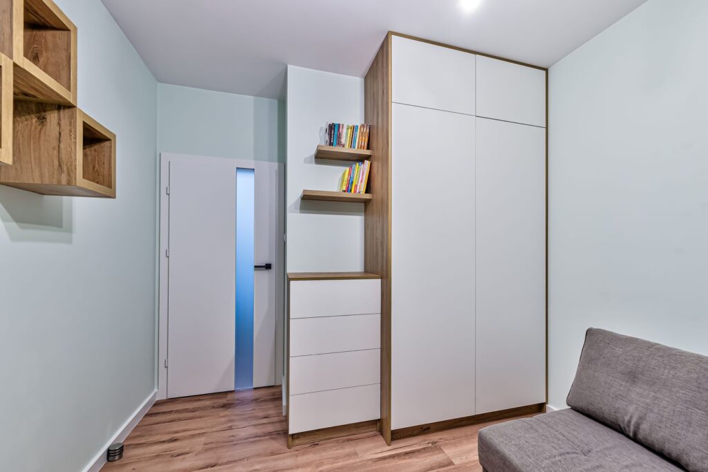 Nowoczesny pokój z jasną szafą, półkami z książkami i szarą sofą na drewnianej podłodze.