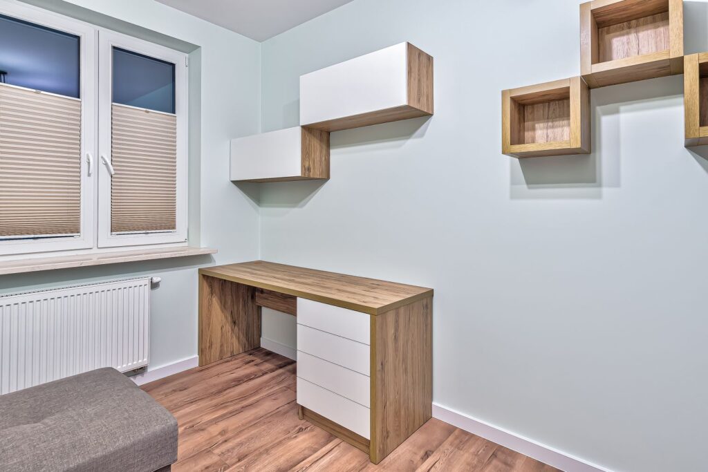 Minimalistyczne biuro z drewnianym biurkiem, białymi półkami i dużym oknem z żaluzjami, jasne ściany.